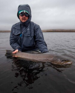 A fisherman displays a large rainbow trout caught in a outdoor freshwater setting. Rainbow trout being held in water by an angler wearing waterproof gear on a cloudy day.