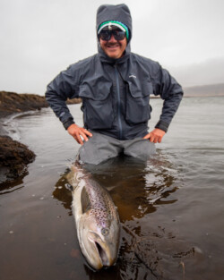 Angler in waterproof gear with sunglasses proudly displays his large brown trout after catching it at the river. Fish partner, fishing, rainbow trout catch, angler with fish, outdoor fishing adventure.