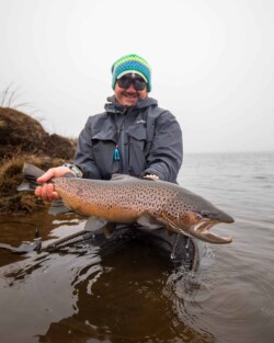 Happy angler holding a large rainbow trout during fishing trip in natural river environment. Vibrant brown trout caught by angler in a foggy lakeside setting, showcasing successful fishing with Fish Partner.