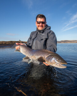 Happy man holding a large rainbow trout in a scenic river landscape. Vibrant rainbow trout caught while fly fishing in clear waters outdoors.