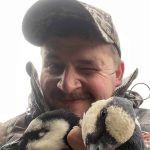 Happy hunter with ducks, enjoying outdoor hunting adventure, wildlife photography, or bird conservation efforts. Young man holding two ducks, outdoors in camouflage gear, smiling and winking, hunting or birdwatching in nature.
