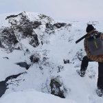 Snow-covered mountain terrain with a hunter in winter gear, carrying a rifle during snow trek. Snowy mountain landscape with a person carrying a rifle, exploring in winter conditions.