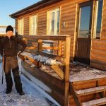 Person standing outside a wooden cabin with snow-covered steps and landscape. Snowy landscape with a wooden cabin in the background, person dressed warmly for winter outdoors.