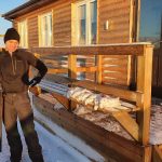 Fishing enthusiast standing outside a cozy cabin with a catch of fish in snowy weather. Freshly caught fish on a snowy porch of a wooden cabin, ready for processing or cooking.